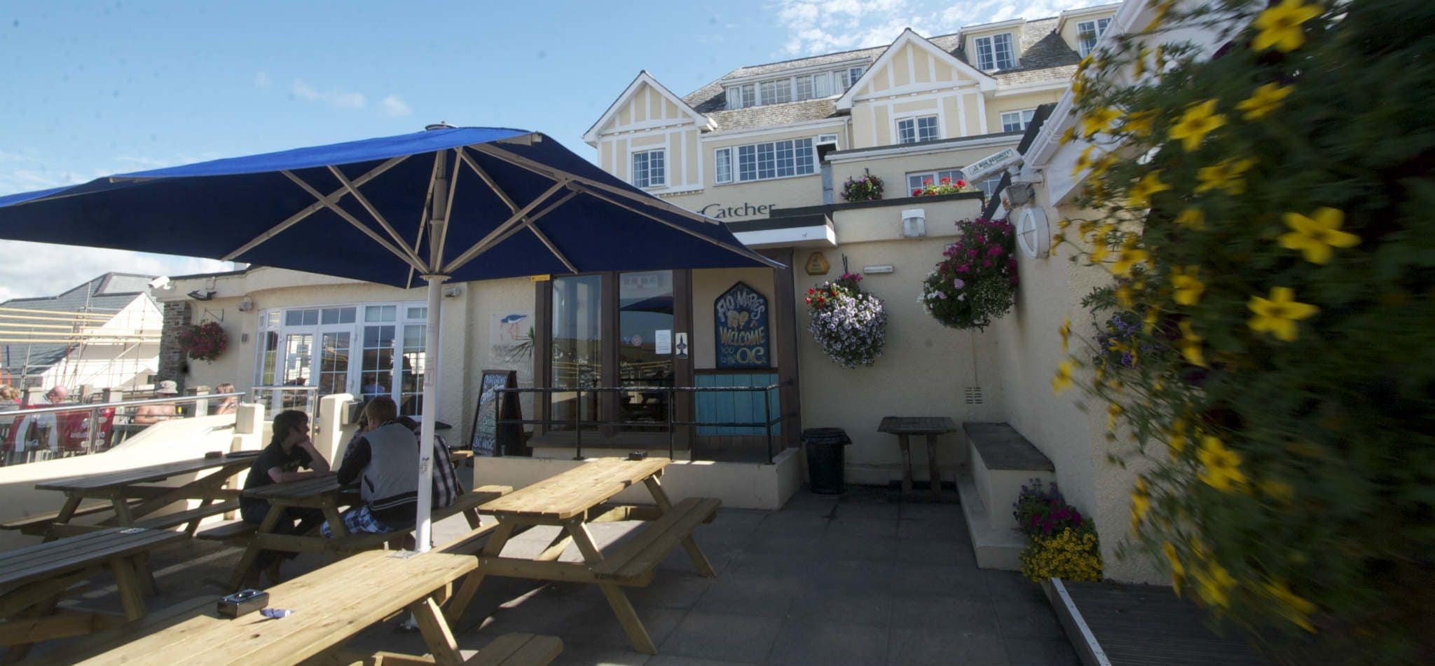 An outdoor seating area with wooden tables and benches, a blue umbrella, and a building with a sign that reads "Catcher".