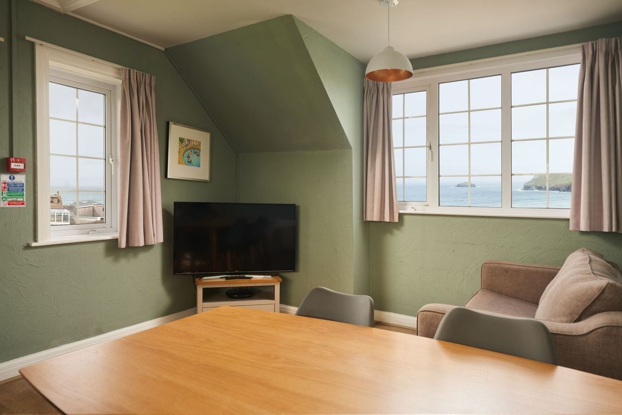 A room with a large wooden table, a television, a sofa, and two windows with curtains.