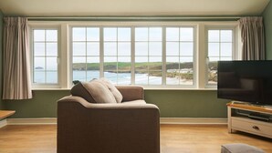 A living room with a large window overlooking a coastal view, a sofa, and a television on a wooden stand. - Oystercatcher Apartments (Wadebridge)