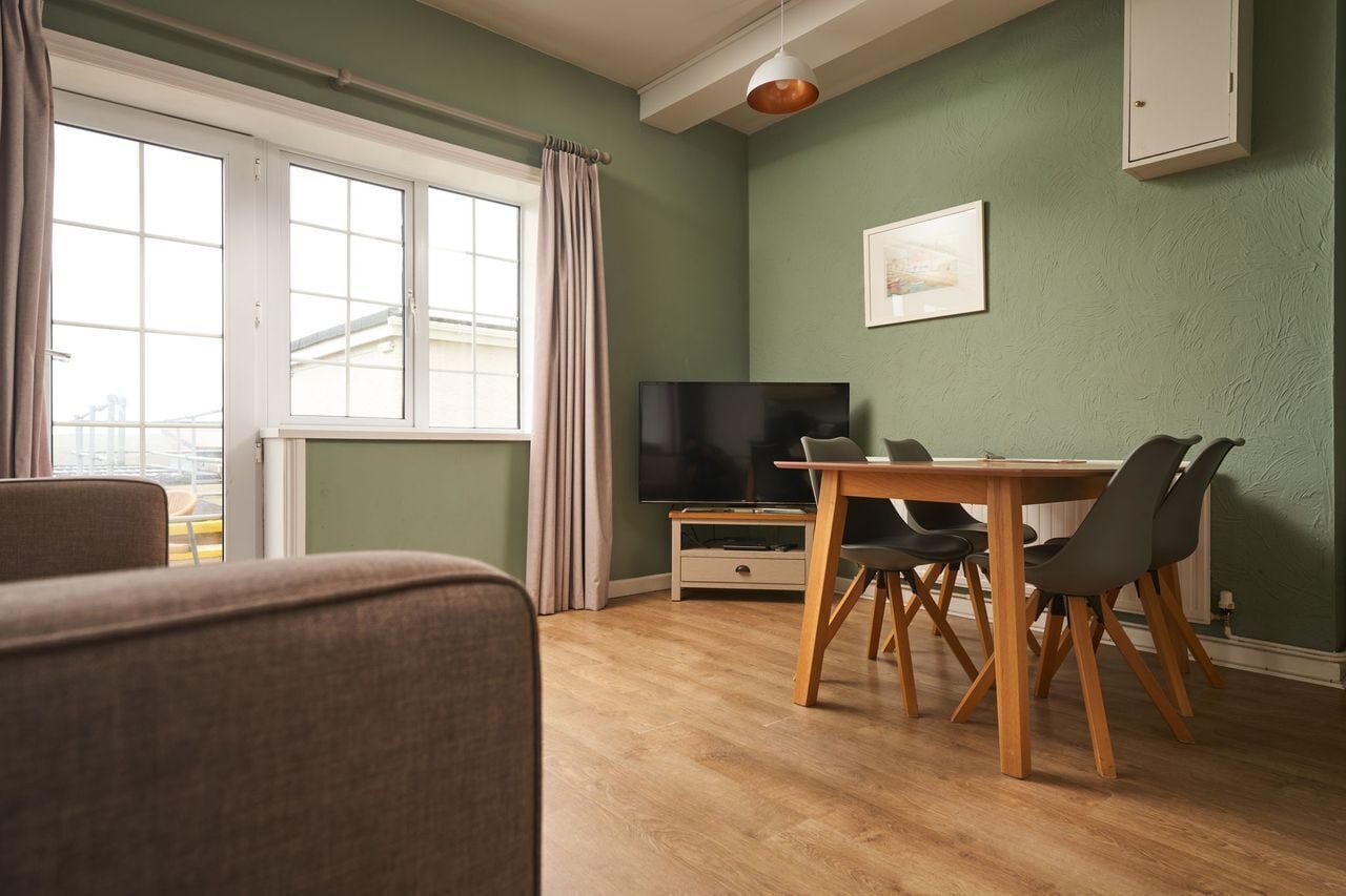 Una sala de estar con televisor, mesa de comedor, sillas y un ventanal con cortinas.