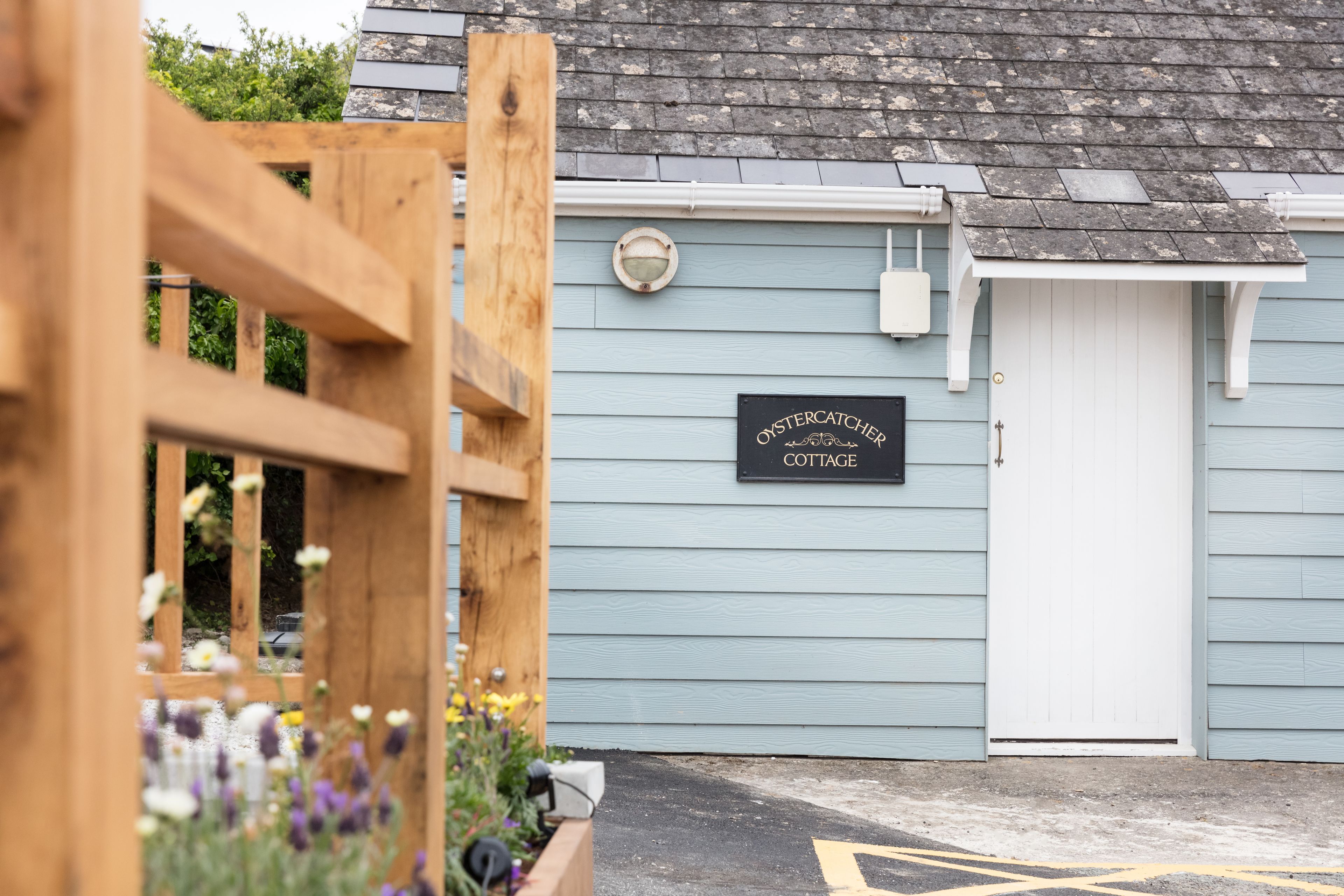 A cottage with a wooden gate, a sign reading "OysterCatcher Cottage," and a white door.