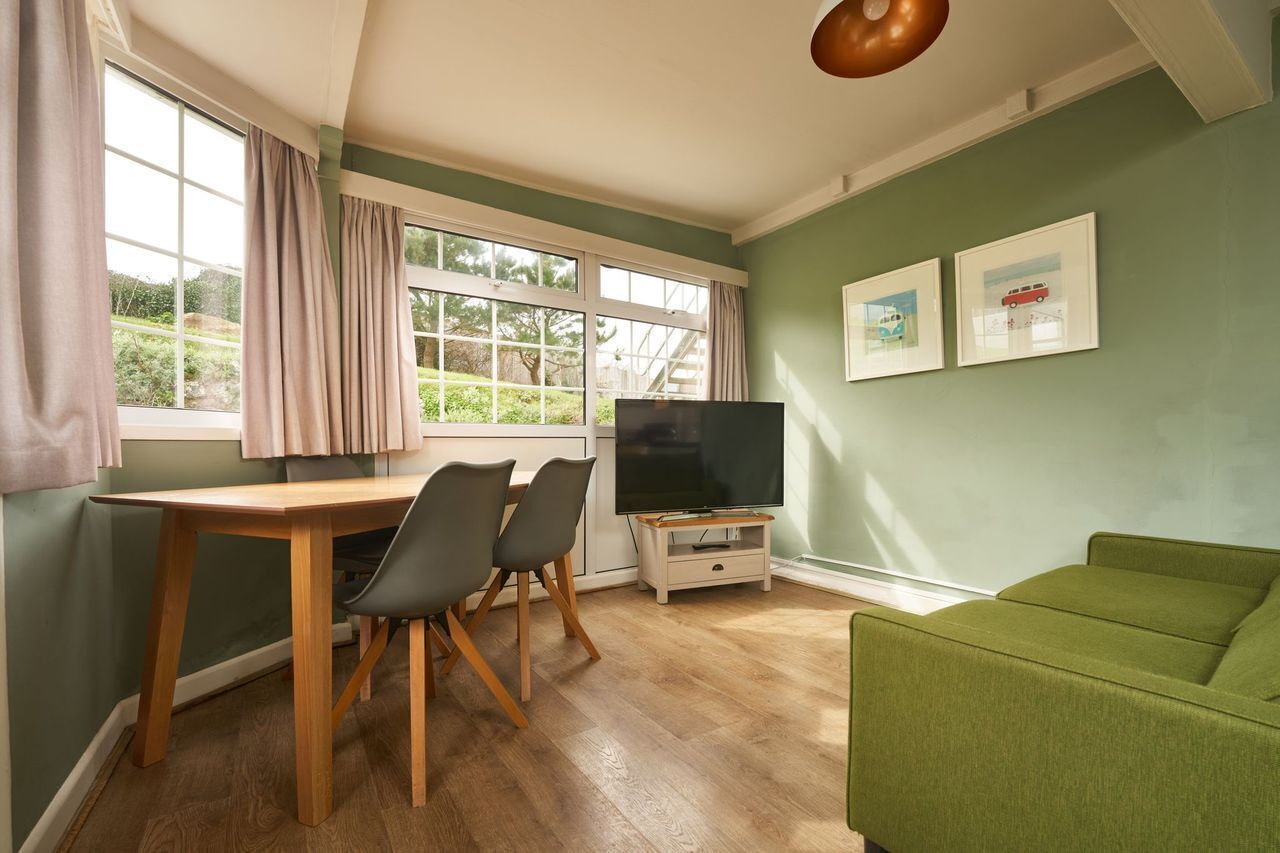 A room with a wooden dining table, two chairs, a television on a stand, a green sofa, and a window with curtains.