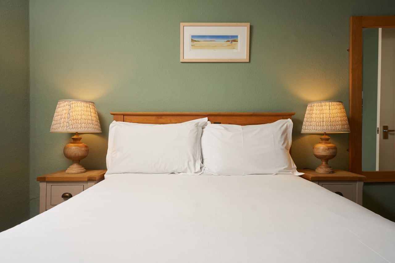 Una cama bien hecha con sábanas blancas, flanqueada por dos mesitas de noche con lámparas, frente a una pared verde con un cuadro enmarcado.