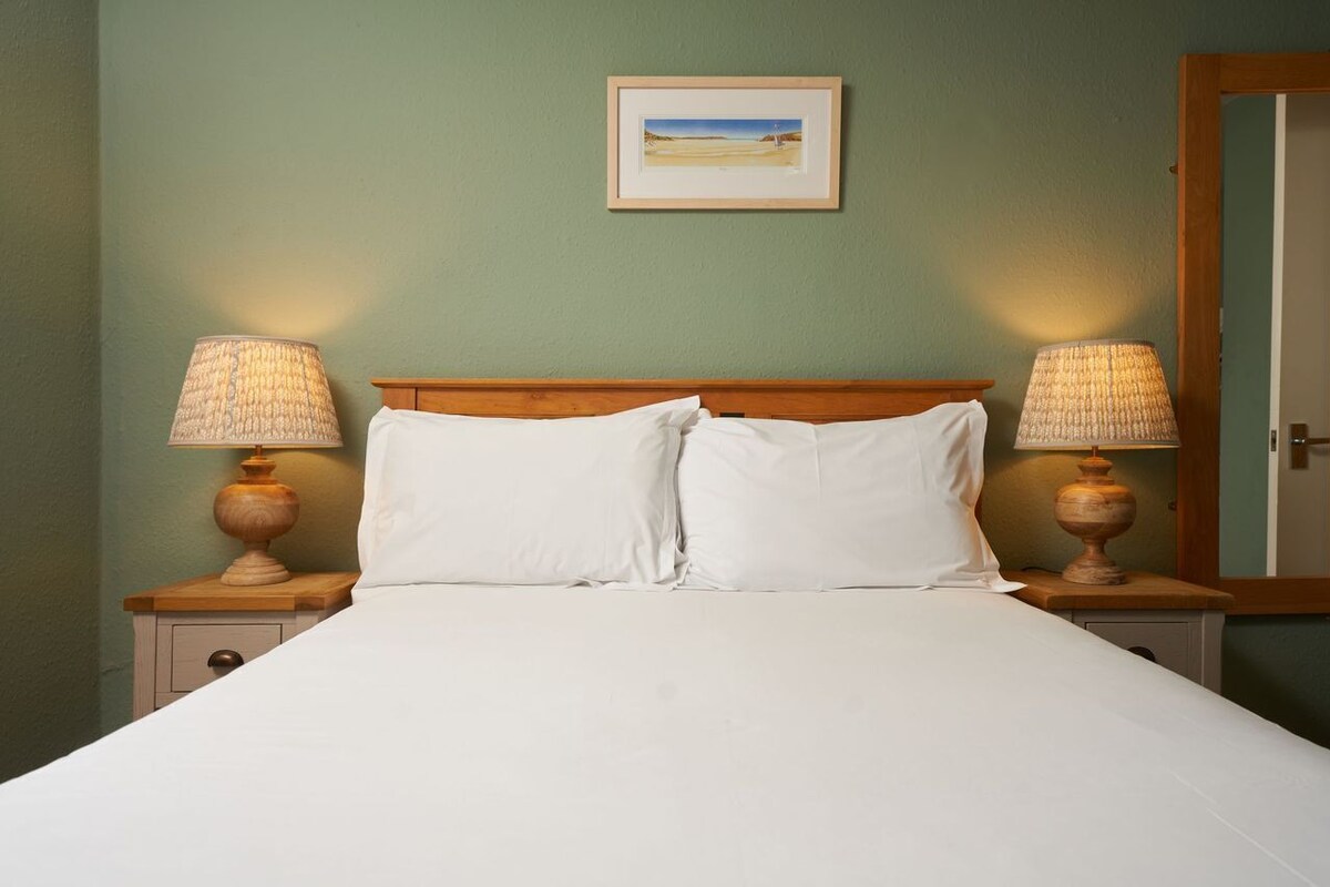 A neatly made bed with white linens, flanked by two bedside tables with lamps, against a green wall with a framed picture above.