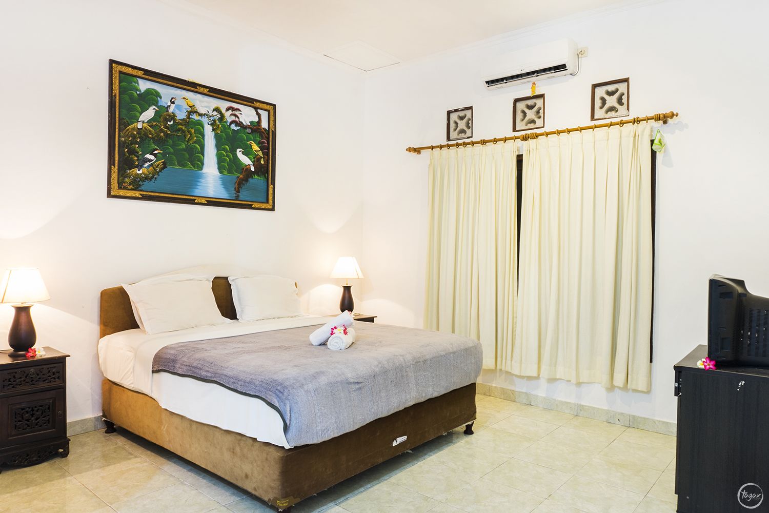 A hotel room with a bed, two bedside lamps, a painting, a curtain, and an air conditioning unit.