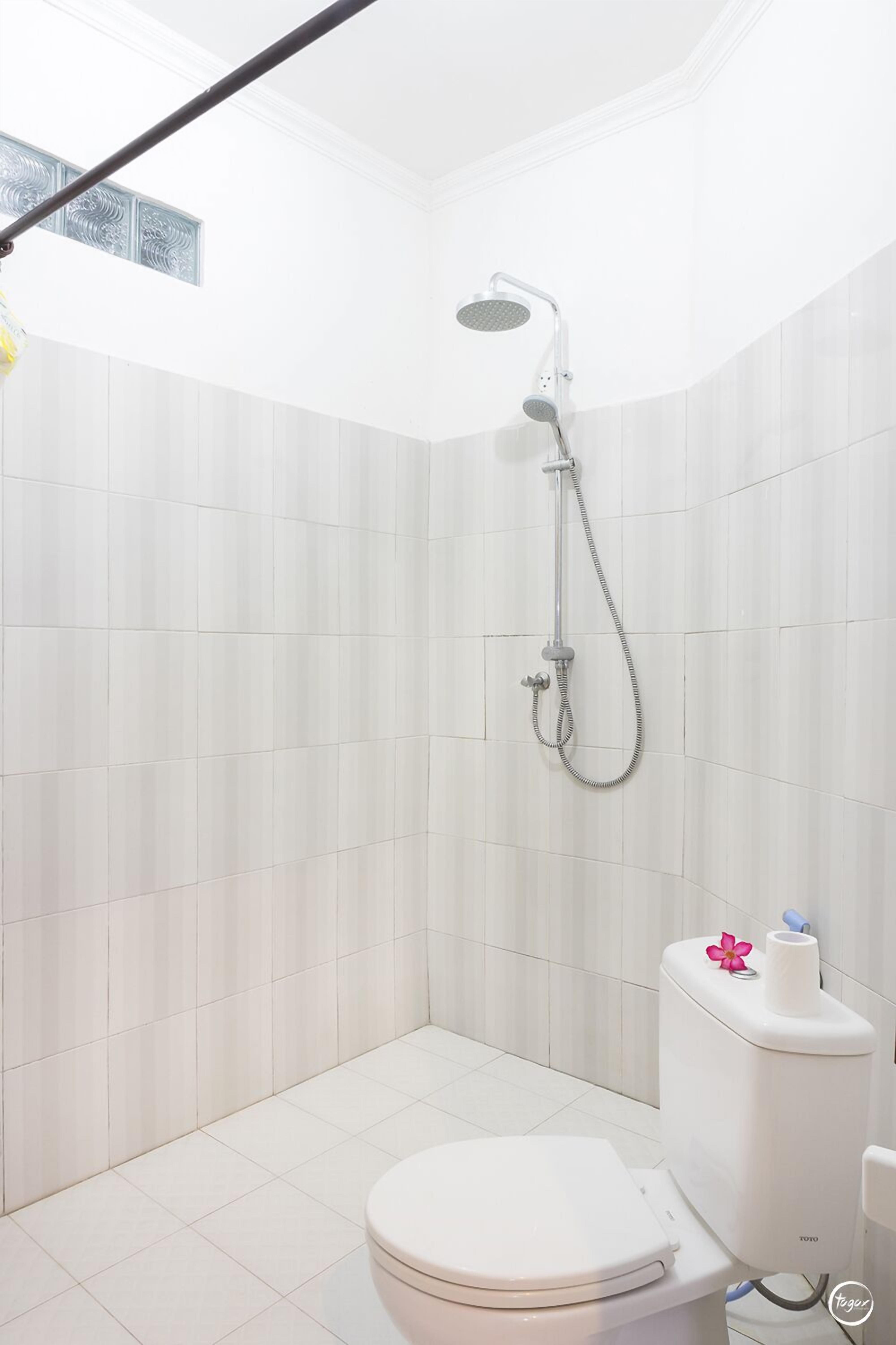 Un baño moderno con azulejos blancos, un inodoro, una ducha con cabezal de ducha manual y un dispensador de papel higiénico con una flor rosa.