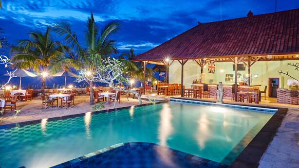 A poolside area with outdoor seating and a bar, surrounded by palm trees and a building with a tiled roof. - Scoobydoo Bungalow (Lembongan Island)