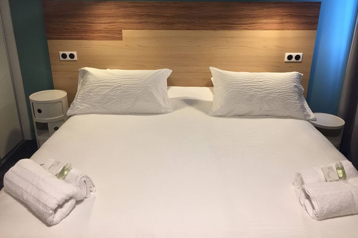 A double bed with white linens, two pillows, two bedside tables, and two rolled towels.