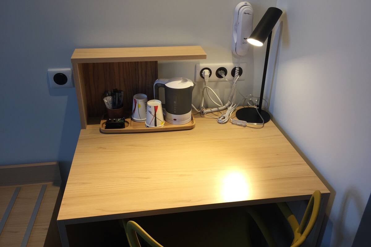 A small, well-lit workspace with a wooden desk, a coffee maker, a lamp, and a power strip.