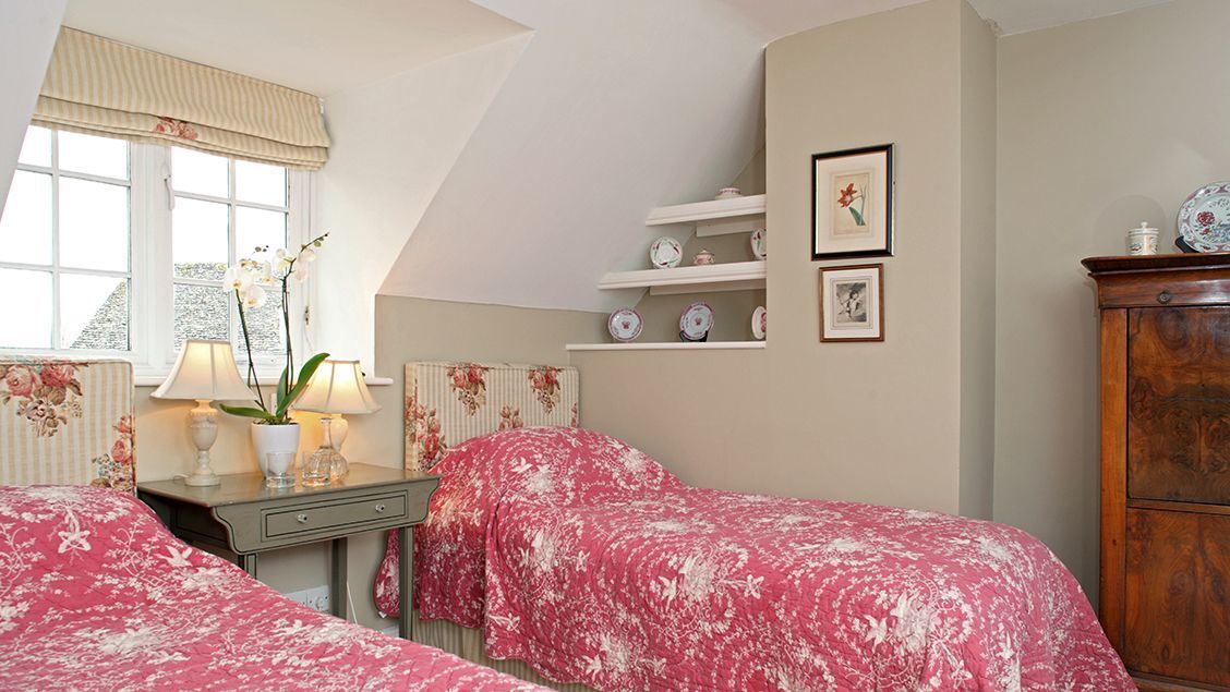Un dormitorio con dos camas, un aparador, una ventana con cortinas y estantes con objetos decorativos.