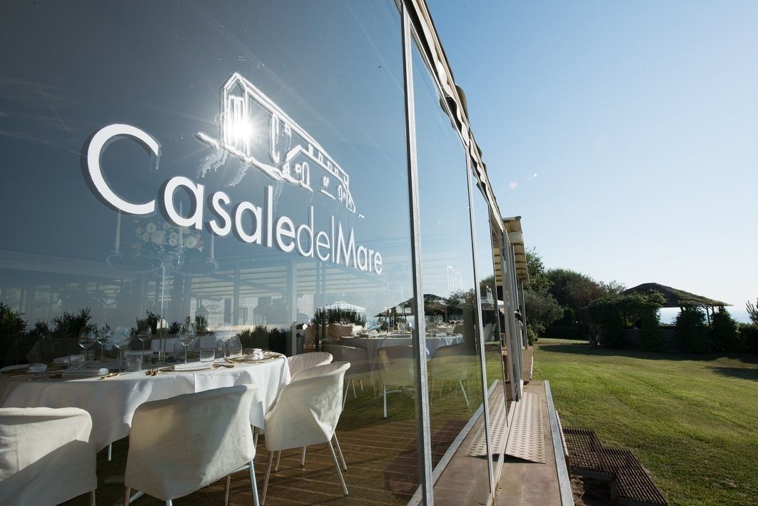 Ein Restaurant mit Außenbereich, einer Glaswand mit der Aufschrift "Casale del Mare" und Blick auf eine Wiese sowie Hügel im Hintergrund.