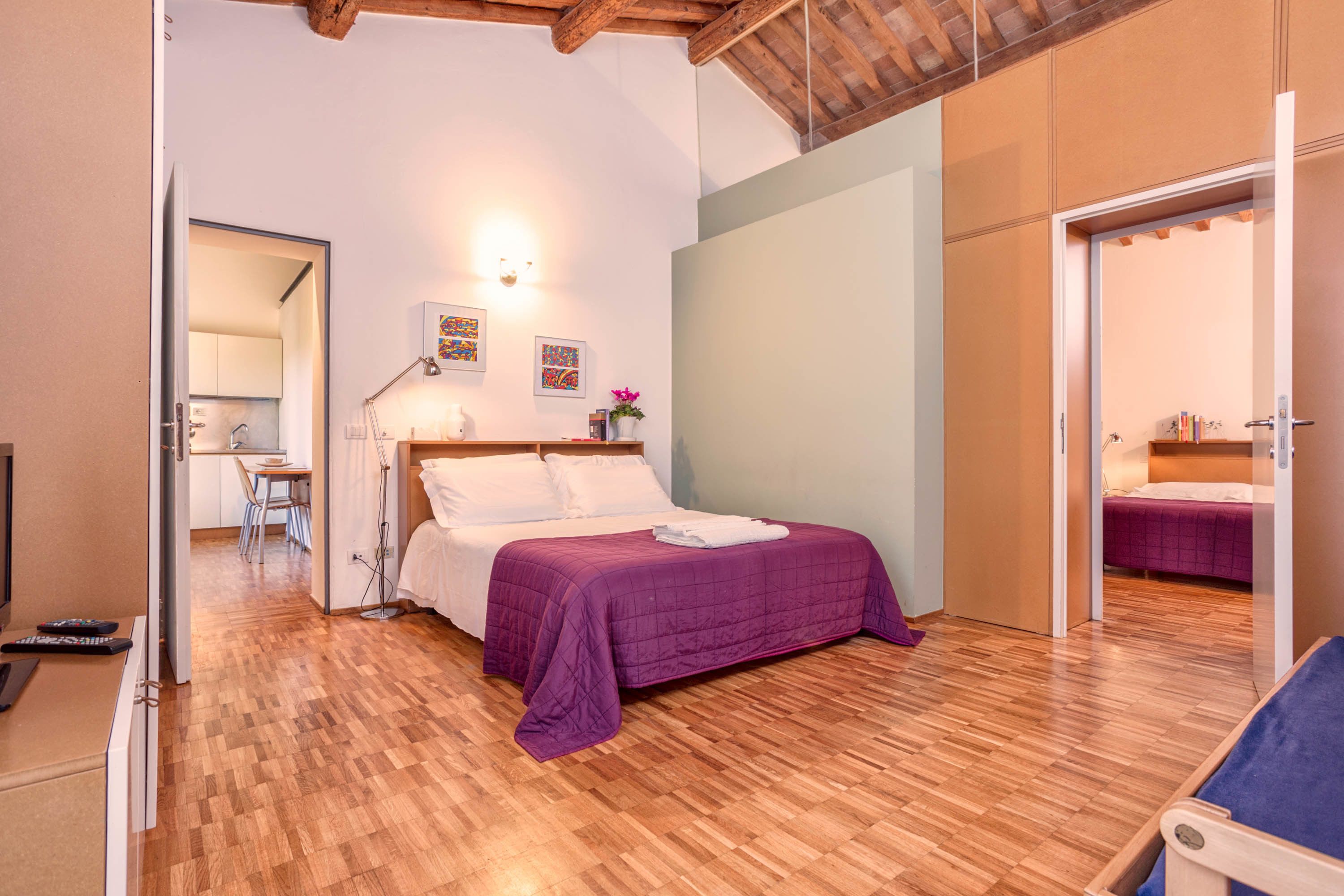 Quarto com piso de madeira, cama com colcha roxa, escrivaninha com cadeira e espelho.
