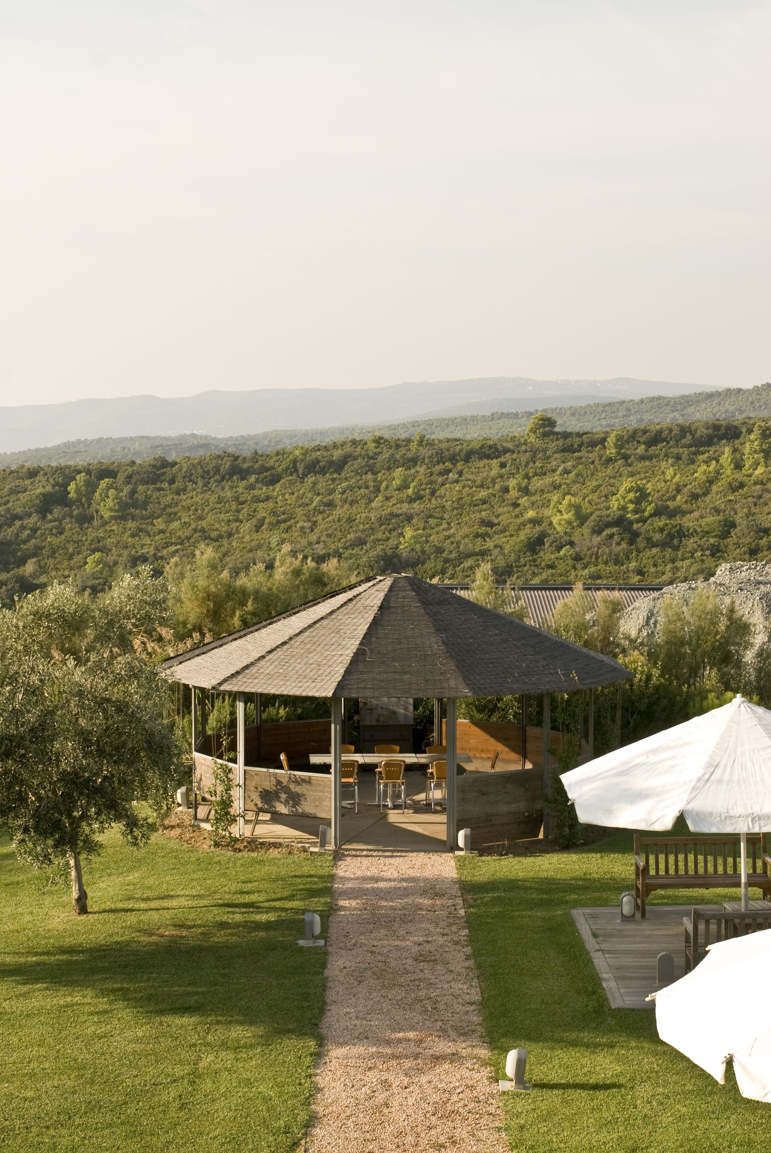 A gazebo with a gravel path leading to it, surrounded by lush greenery and a view of distant hills.