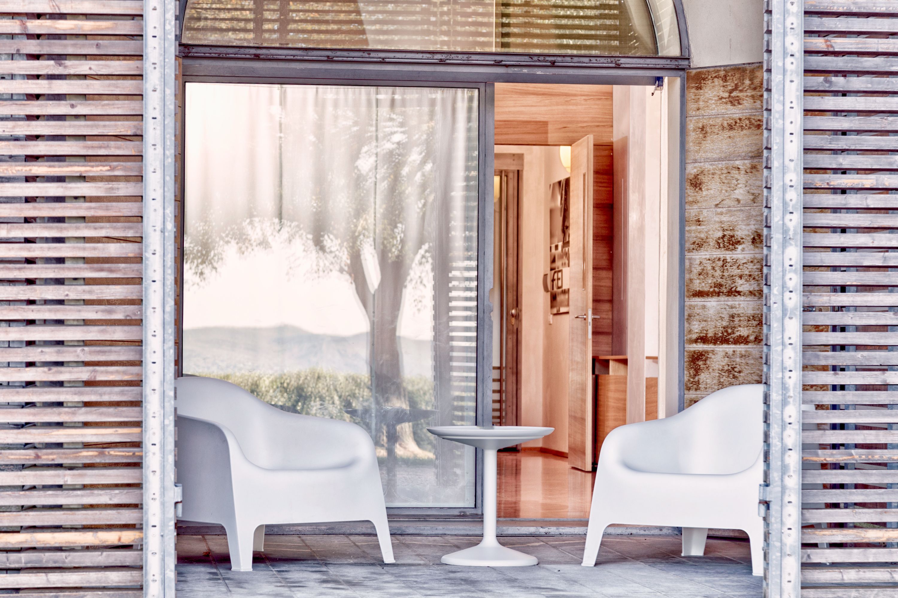 A modern interior with two white chairs and a small table, a wooden door, and a view of a tree and mountains through a large window.