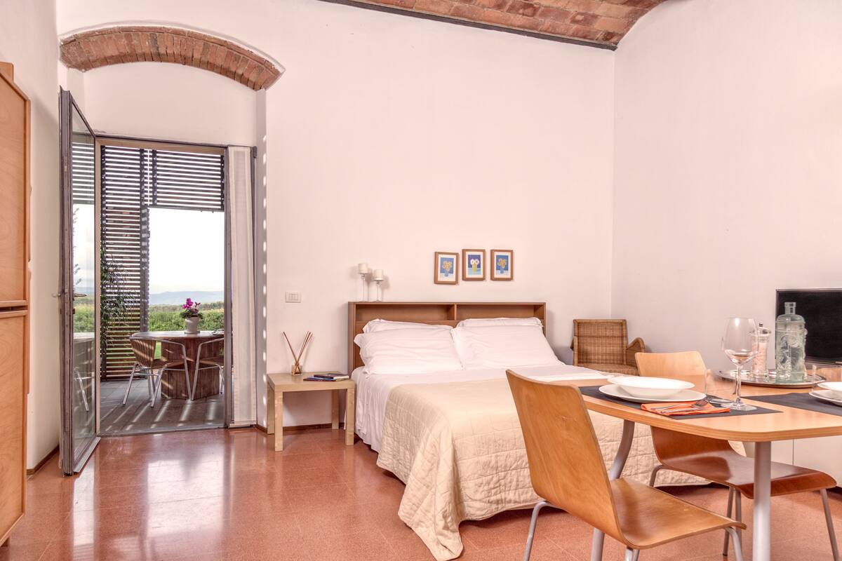 A bedroom with a bed, a dining table, and a balcony with a table and chairs.