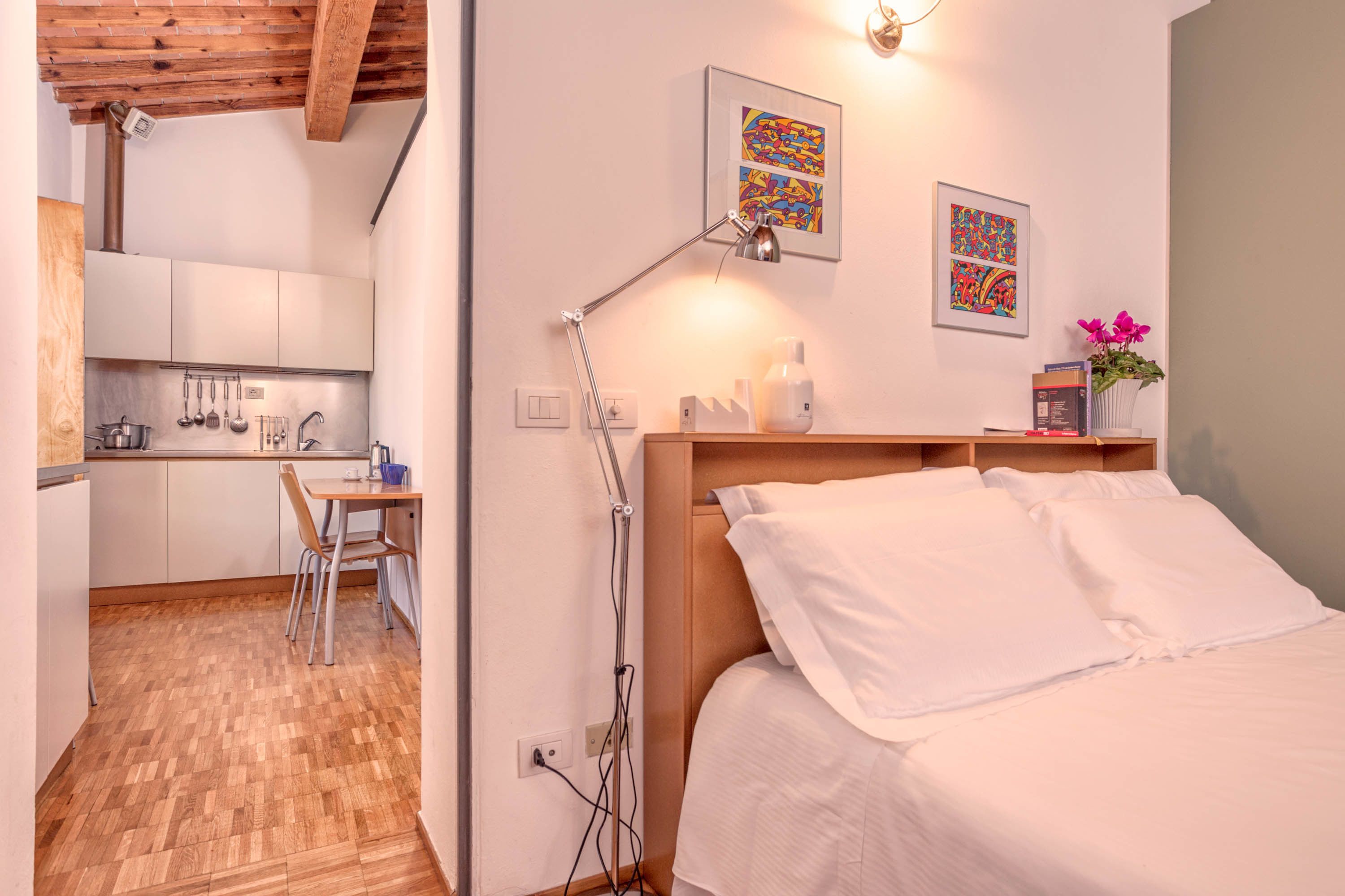 Habitación de hotel compacta con techo de madera, cama con ropa blanca, escritorio con silla y una kitchenette al fondo.