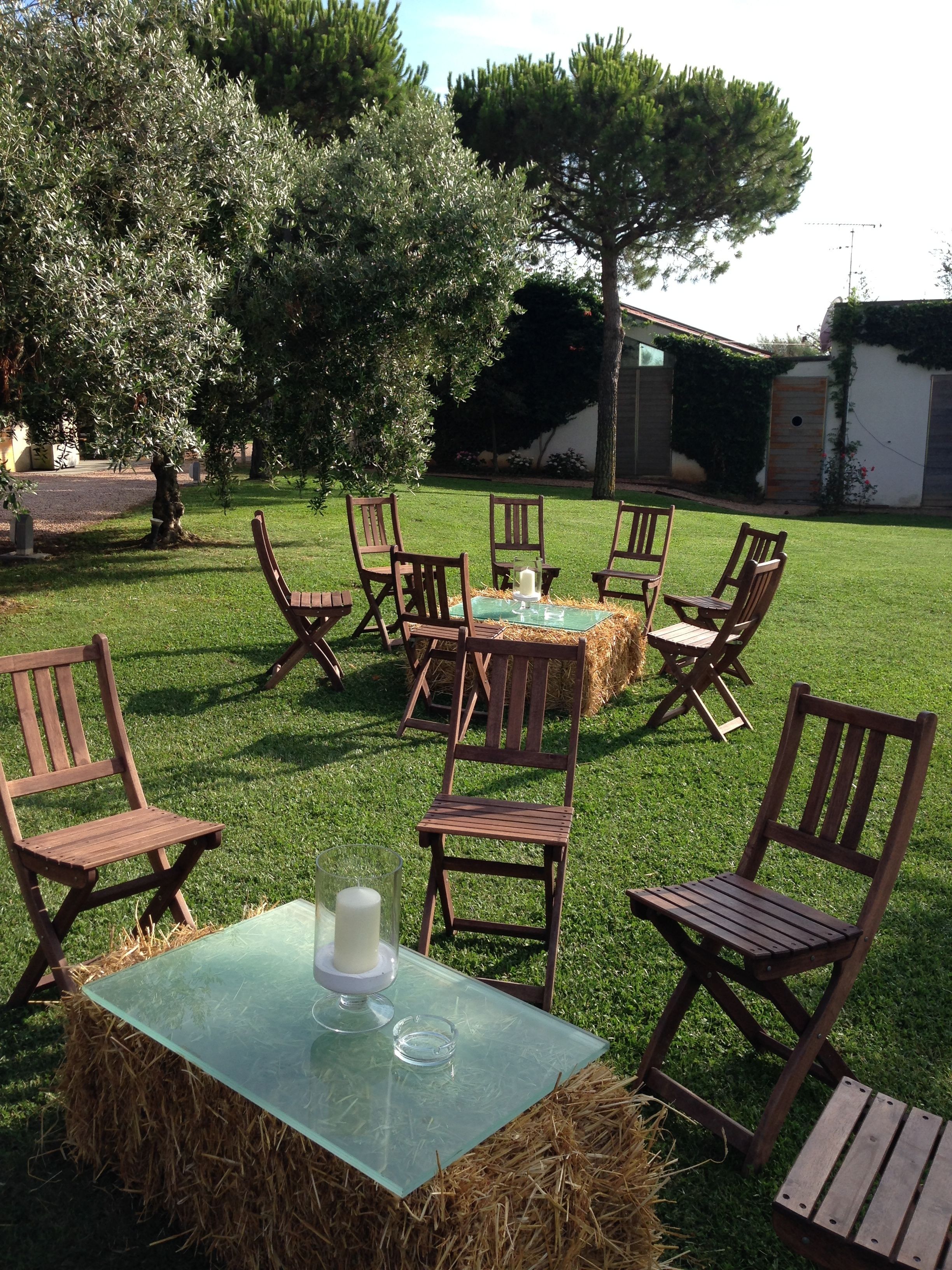 Un espacio de estar al aire libre con una mesa de vidrio rodeada de sillas de madera, fardos de heno y árboles.