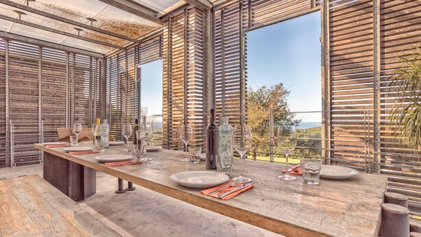 A long dining table with multiple plates, glasses, and cutlery, set against a backdrop of wooden slats and large windows overlooking a scenic view.