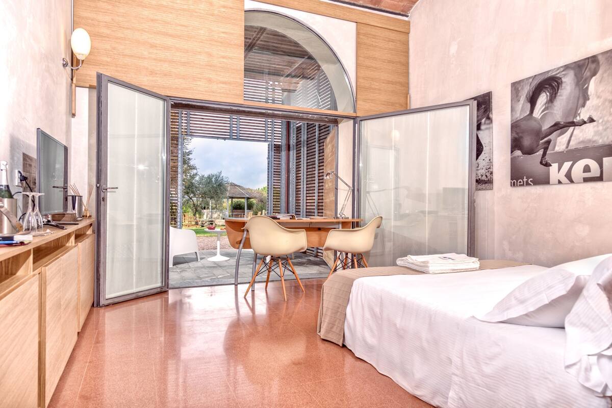 A modern bedroom with a bed, a desk, and chairs, a glass door leading to an outdoor area, and a poster on the wall.