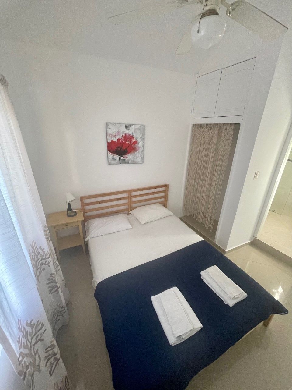 A bedroom with a bed, a nightstand, a lamp, a painting, and a ceiling fan.