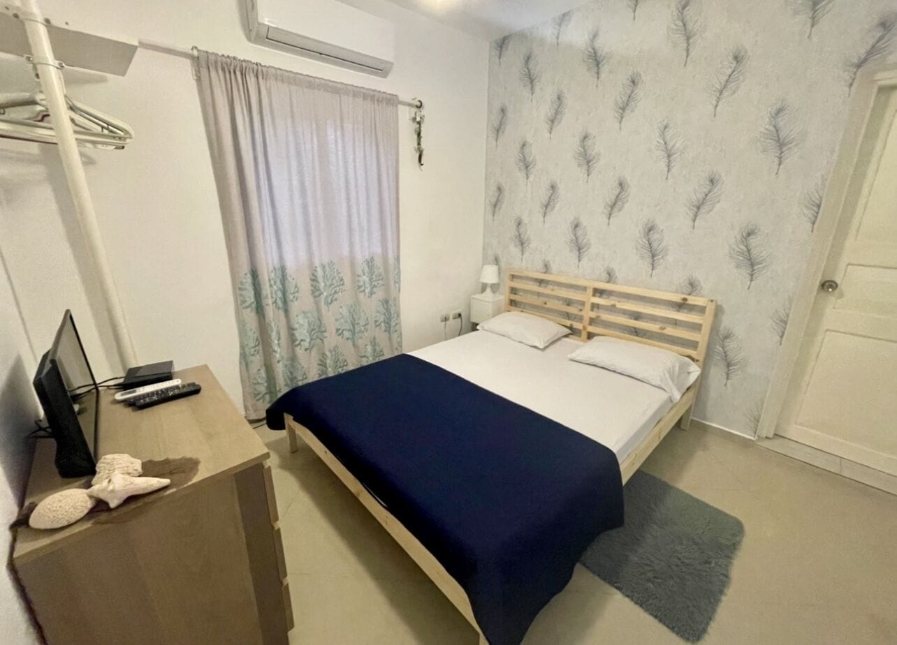 A bedroom with a bed, a desk with a computer and remote controls, a window with curtains, and a wall with a feather pattern.