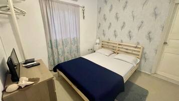 A bedroom with a bed, a desk with a computer and remote controls, a window with curtains, and a wall with a feather pattern.