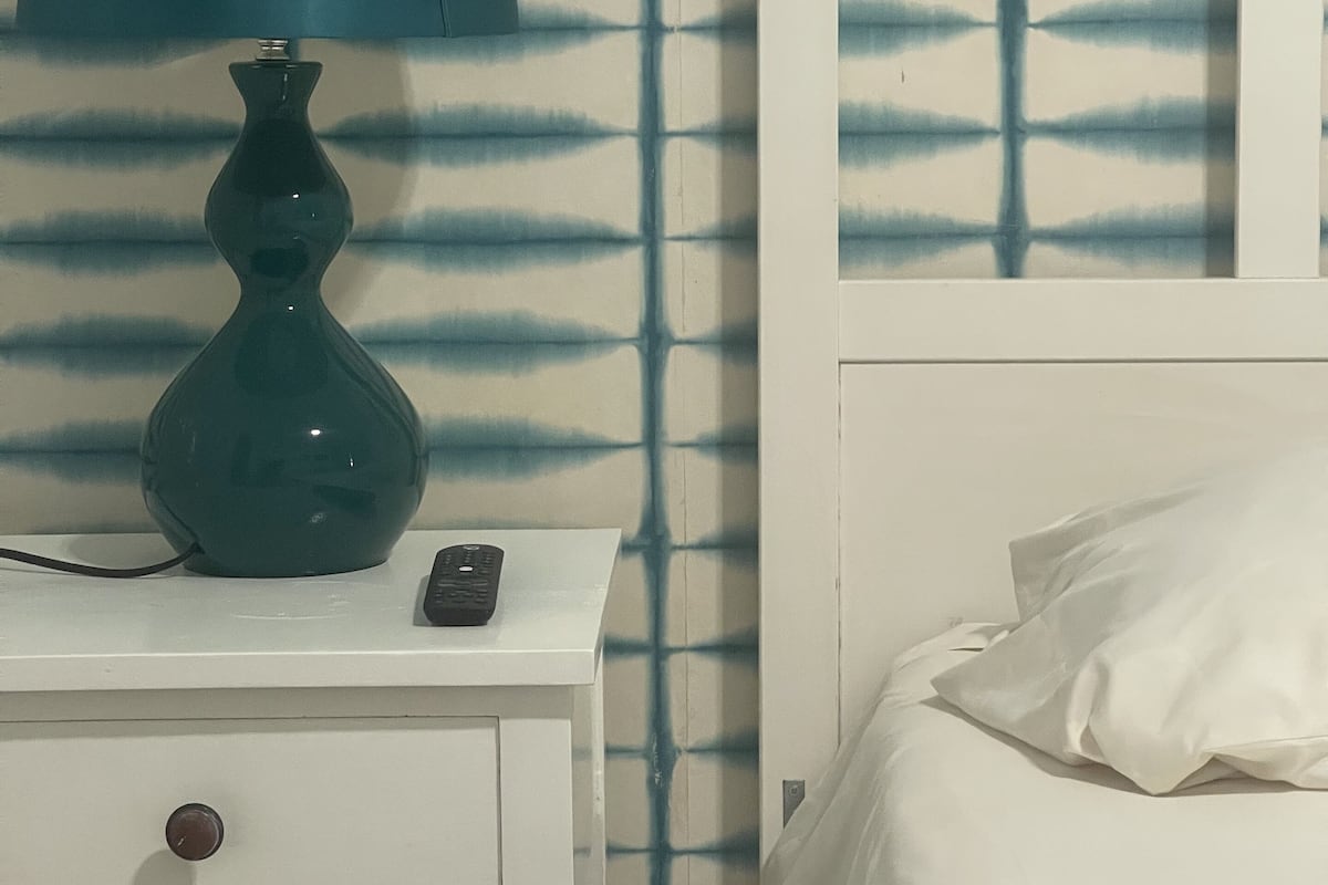 Une table de chevet blanche, sur laquelle se trouvent une lampe turquoise, une télécommande et un tiroir. On aperçoit un lit blanc avec un oreiller blanc et une parure de lit à rayures.