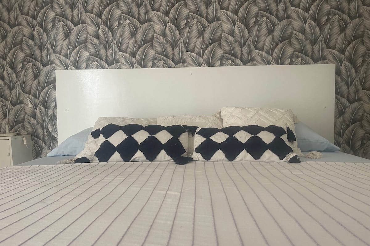 Un lit avec une couvre-lit à rayures, deux oreillers à motifs losangés et une tête de lit placée devant un papier peint à motifs de feuilles.
