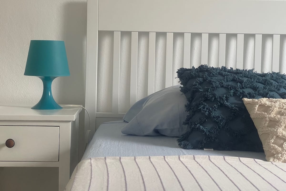 Un lit bien fait, recouvert d’une couvre-lit à rayures, une lampe de chevet de couleur turquoise et une table de chevet blanche avec un tiroir.