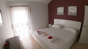 Ein ordentlich bezogenes Bett mit weißen Leinen und einer roten Blume, ein Nachttisch mit Lampe und ein Fenster mit Vorhängen.