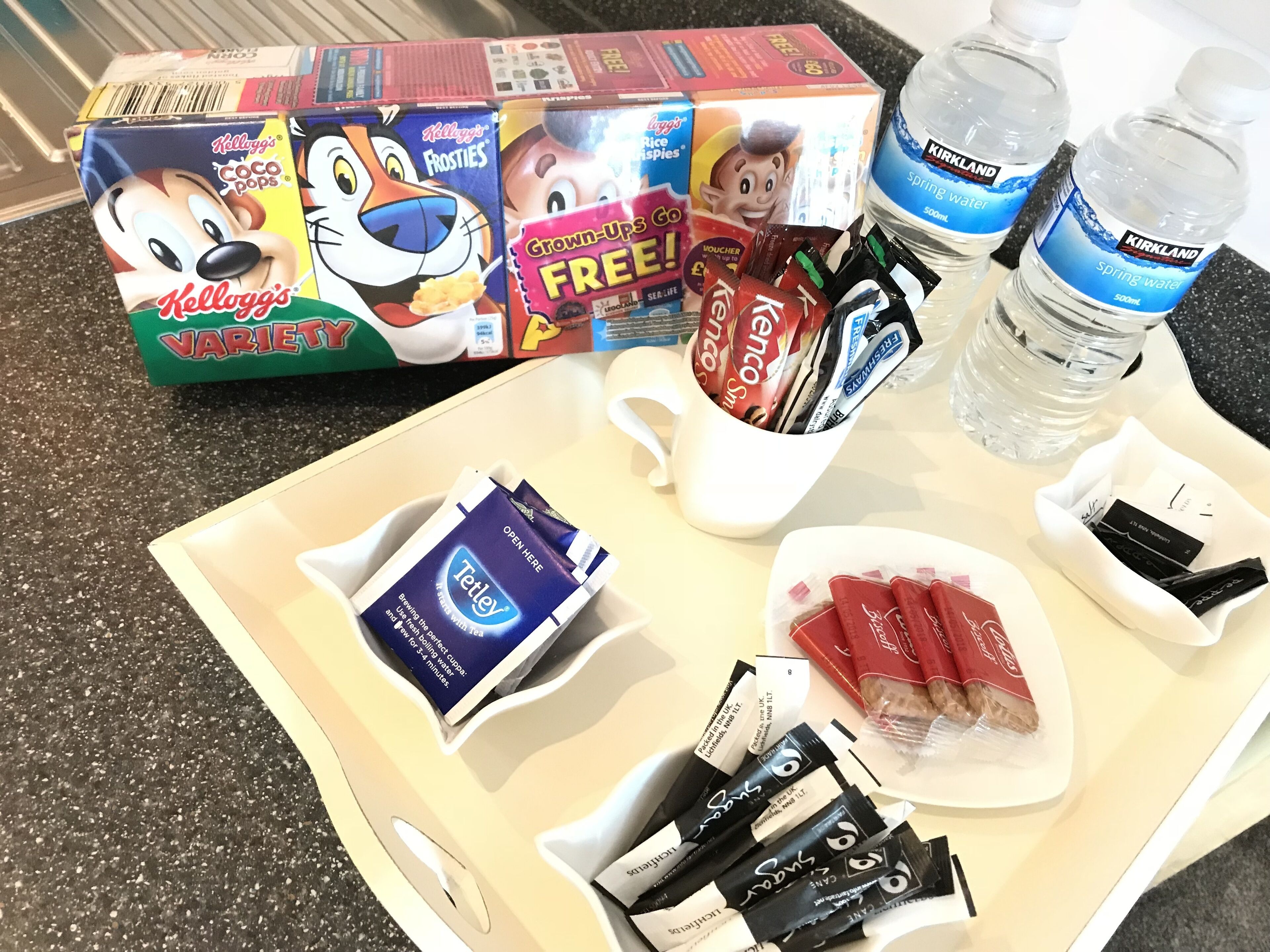 En bakke med forskellige spiser, herunder cornflakes, vand på flaske og snacks.