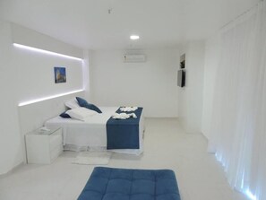 A modern bedroom with a bed, bedside table, and a blue ottoman. - Hotel Real (Cabo Frio)