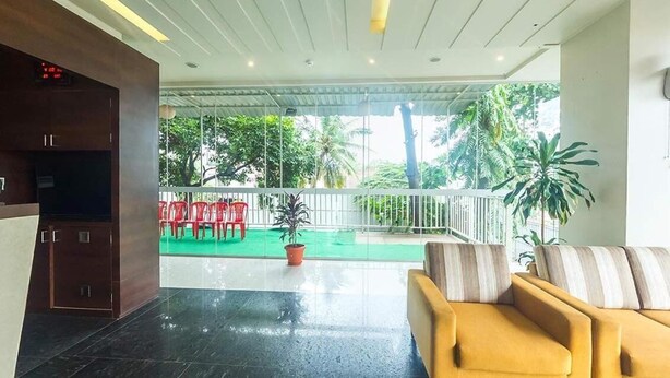 A lobby with a yellow sofa, a TV, a green outdoor area with red chairs, and a potted plant. - Hotel Rasika (Kolhapur)