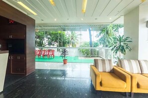 Un vestíbulo con un sofá amarillo, televisor, zona exterior verde con sillas rojas y una planta en maceta.