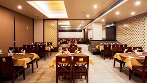 A dining area with multiple tables and chairs, arranged in rows, with a high ceiling and recessed lighting. - Hotel Rasika (Kolhapur)