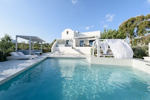 A modern poolside area with a clear pool, white lounge chairs, and a white building with a blue door. - Ambeli luxury villa (Santorini)
