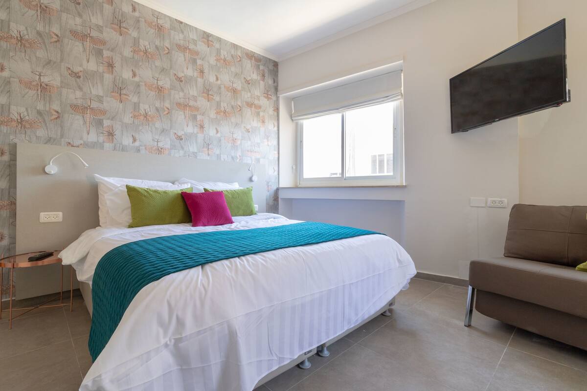 A hotel room with a bed, a TV mounted on the wall, a window, and a small table with a lamp.