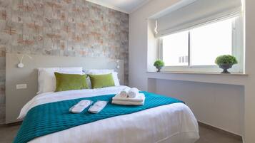 Una cama bien hecha con sábanas blancas, una colcha turquesa y dos almohadas verdes. Se ve una ventana con vistas al exterior y una pequeña planta en maceta en el alféizar.