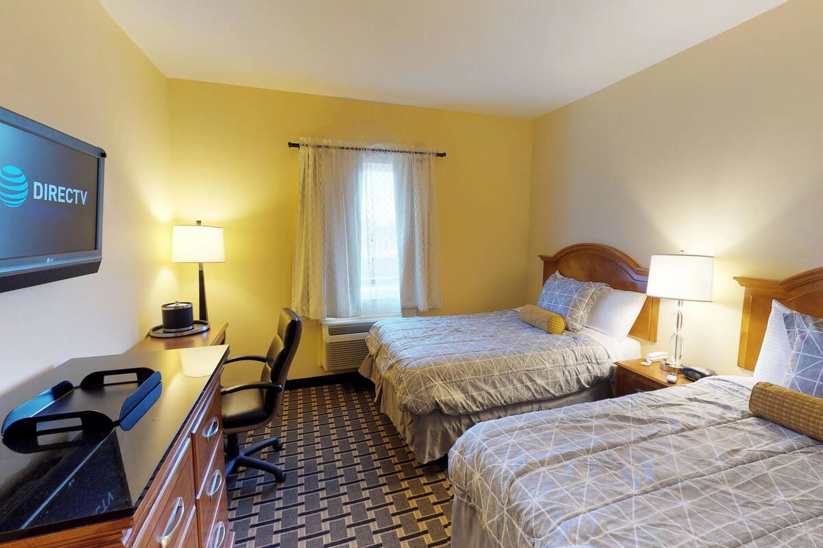 A hotel room with two beds, a desk with a computer, a TV displaying DIRECTV, and a window with curtains.