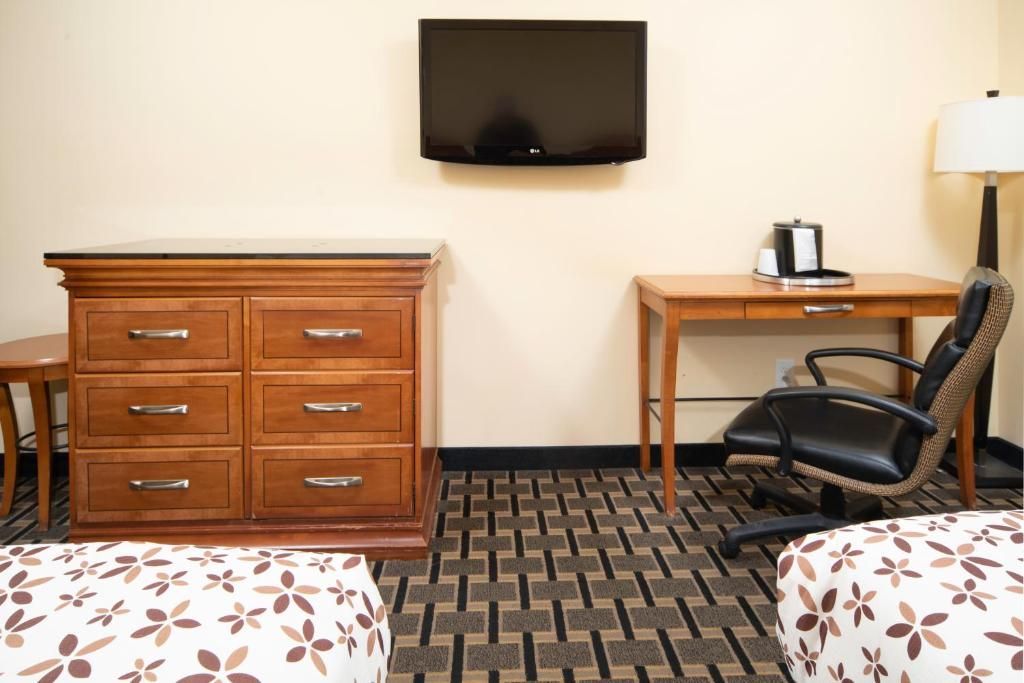 A hotel room with a wooden dresser, a desk with a chair, a television, and a patterned bedspread.