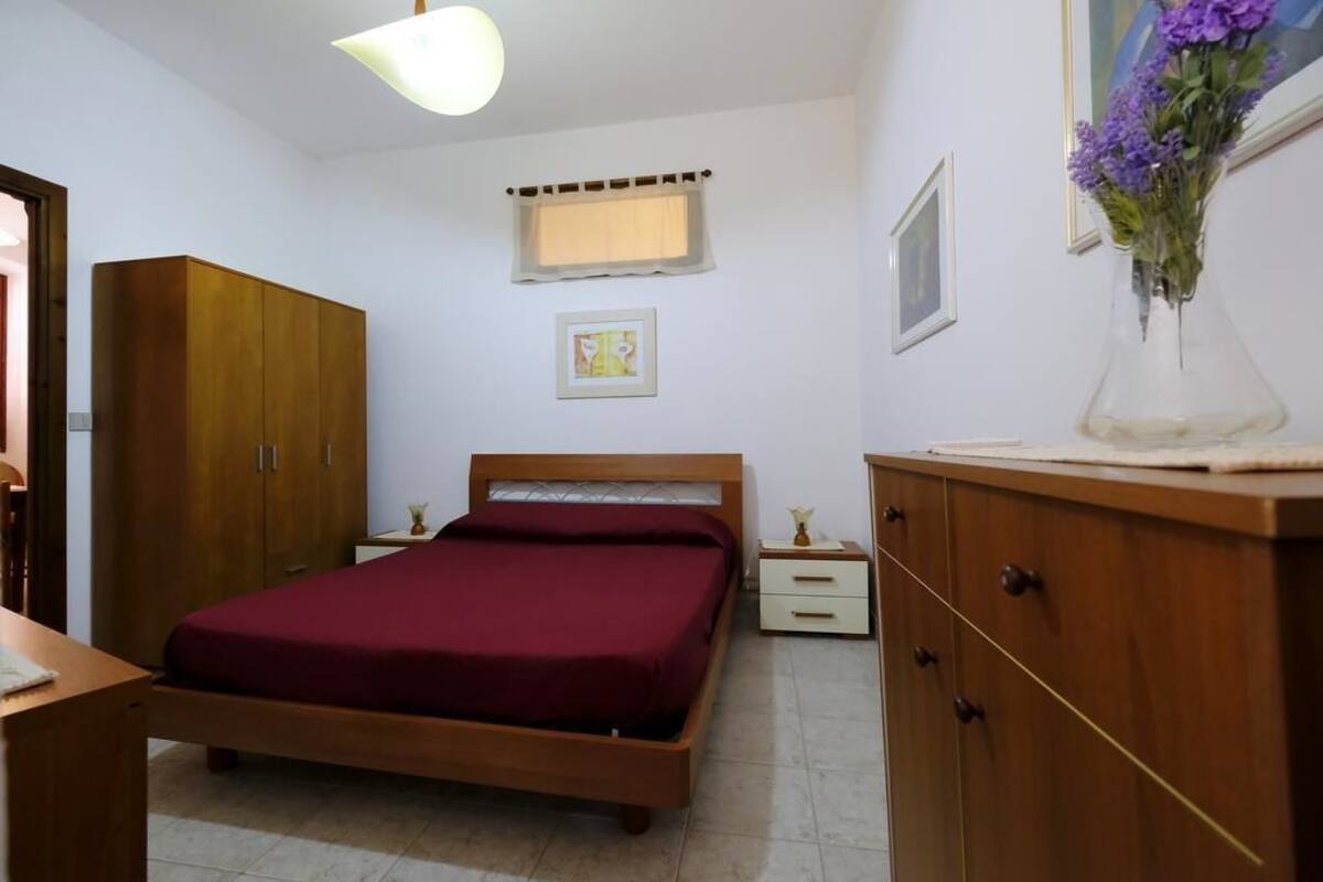 Un dormitorio con cama de madera, una mesita de noche, un aparador, una ventana con cortina y un florero con flores sobre el aparador.