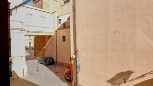 A narrow street with a tower in the background, a red car parked on the side, and potted plants on the balcony railing. - Bed & Breakfast L'aquilino (Iglesias)