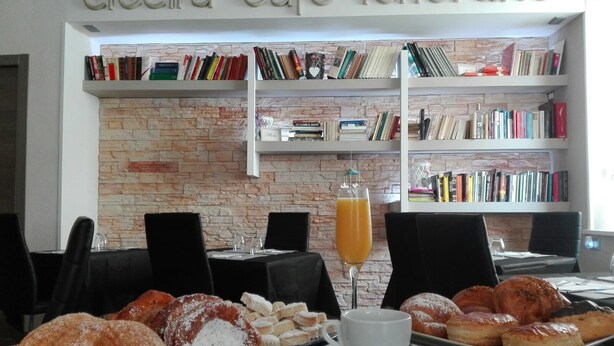 A cafe with a brick wall and bookshelves filled with books. There is a table with pastries, a cup, and a glass of orange juice. - Bed & Breakfast L'aquilino (Iglesias)