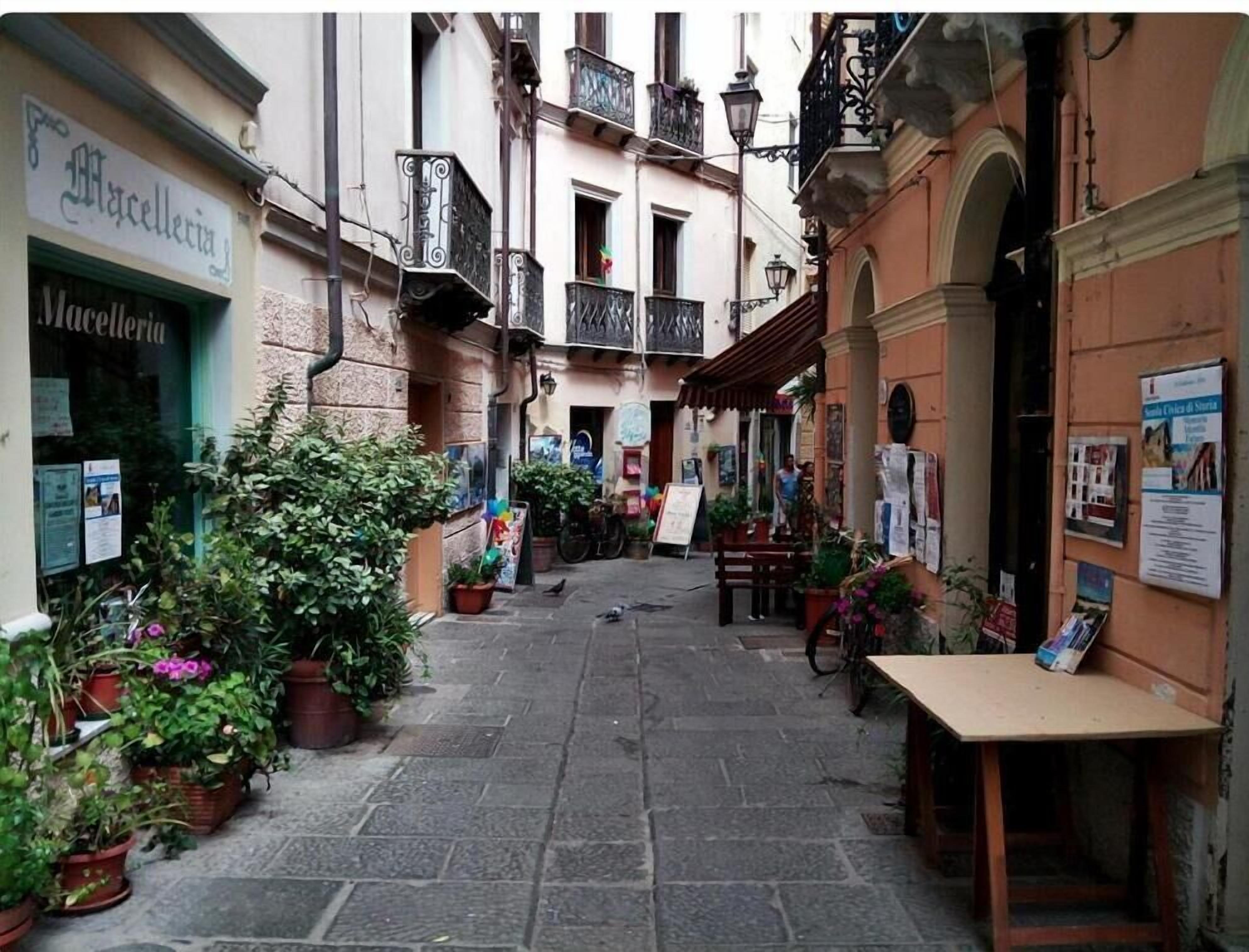 A narrow street with buildings on either side, some with balconies and signage. There are potted plants along the street and a bicycle parked on the right.