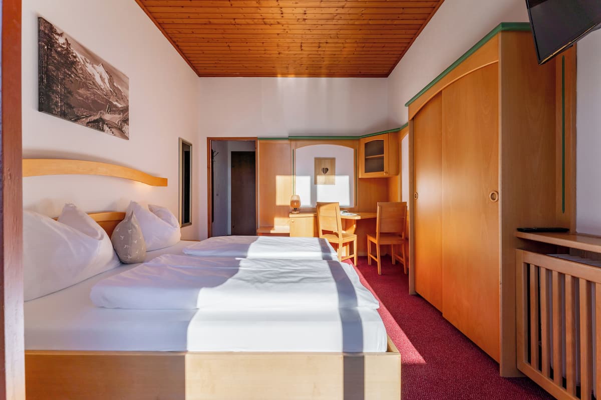 A hotel room with a wooden ceiling, a bed with white linens, a desk with a chair, and a TV mounted on the wall.