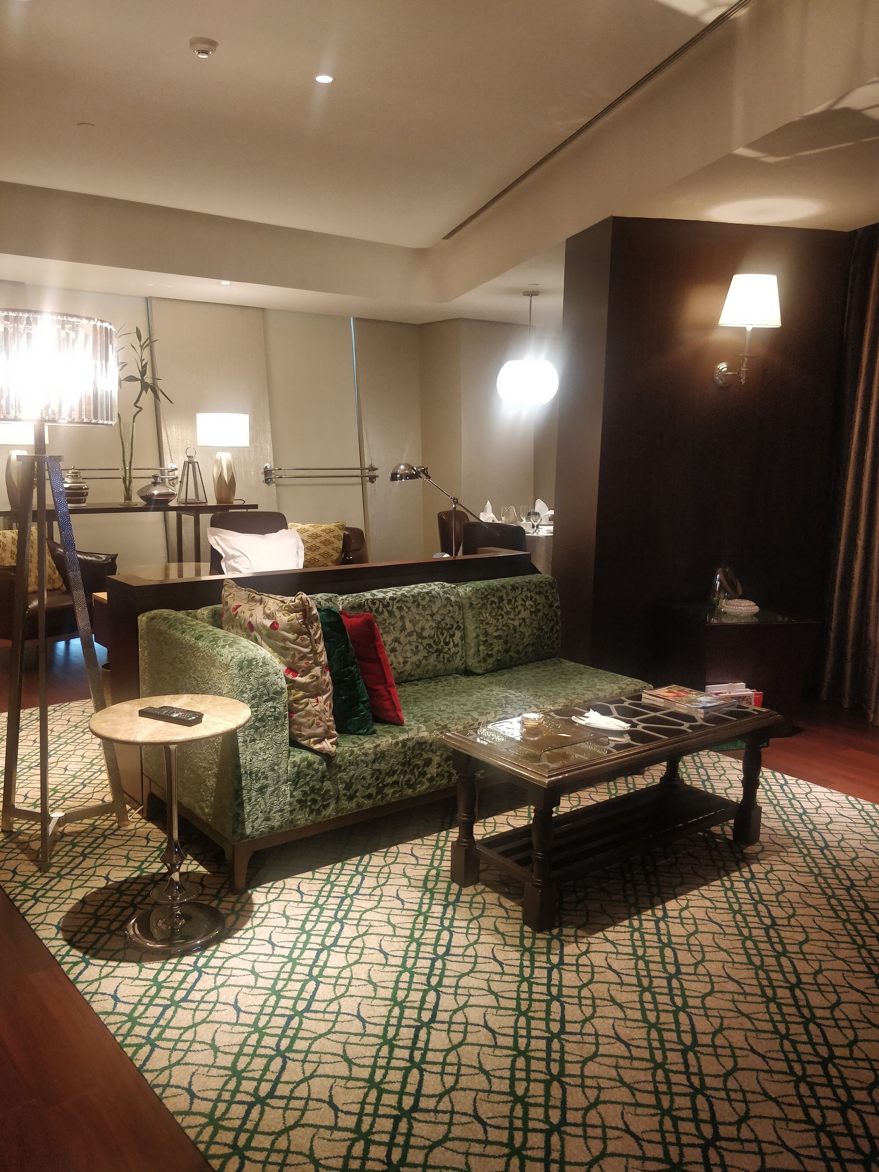 A modern hotel room with a green patterned sofa, a round side table, a coffee table, and a dining area with a table and chairs.