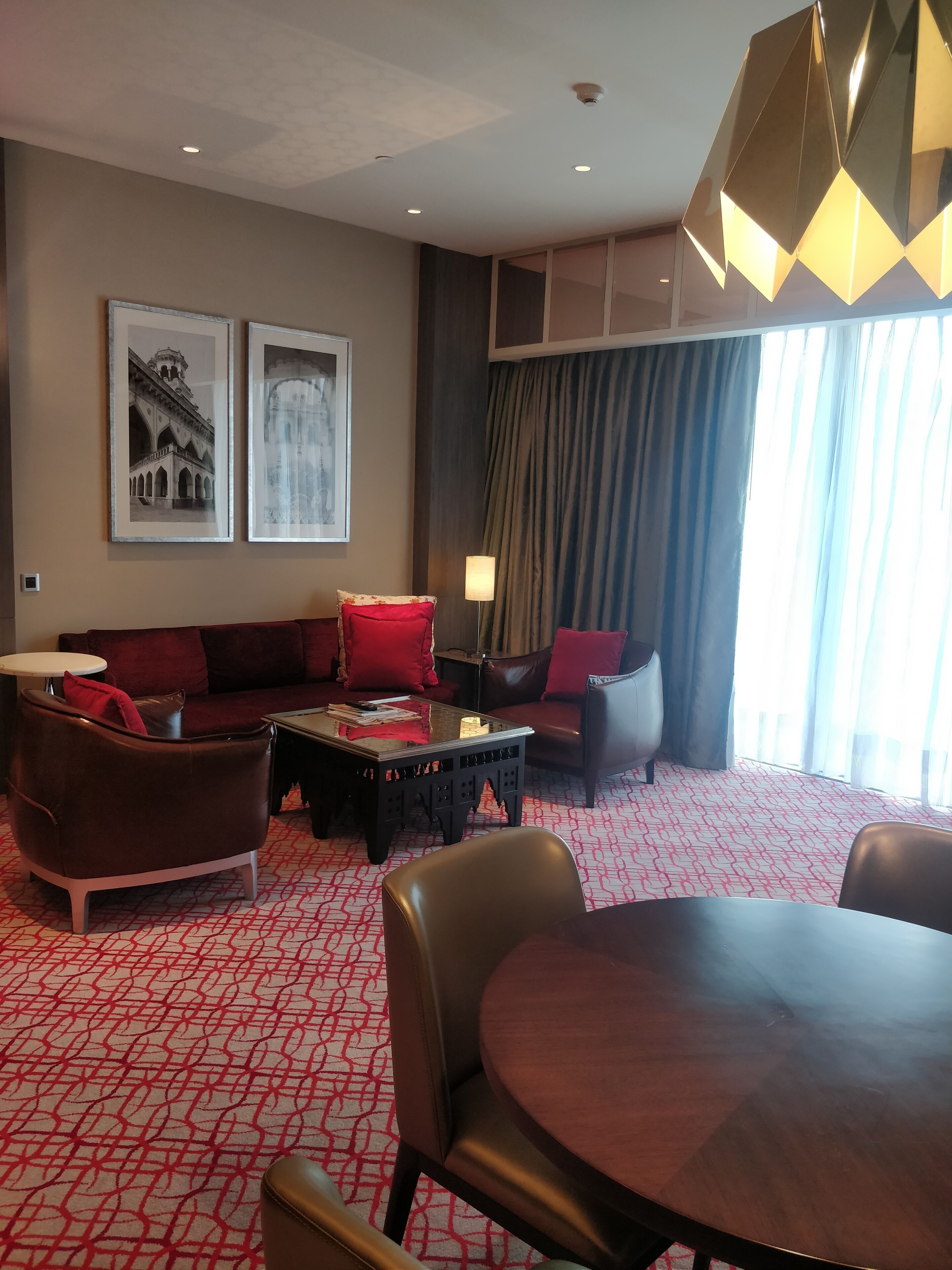A hotel room with a red patterned carpet, a sofa set, a dining table, and two framed pictures on the wall.