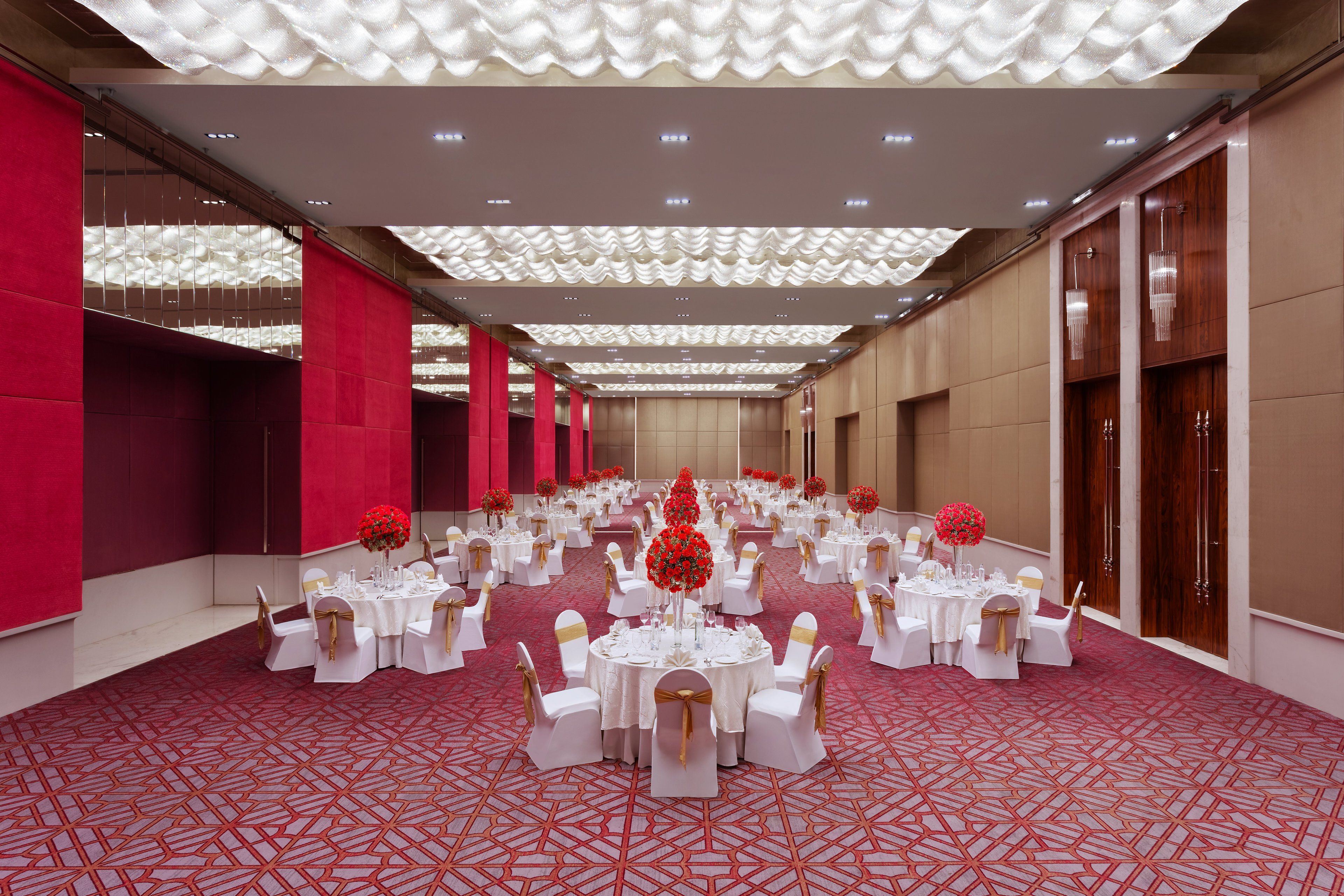Un salón de banquetes con mesas redondas, manteles blancos y centros de mesa florales rojos.