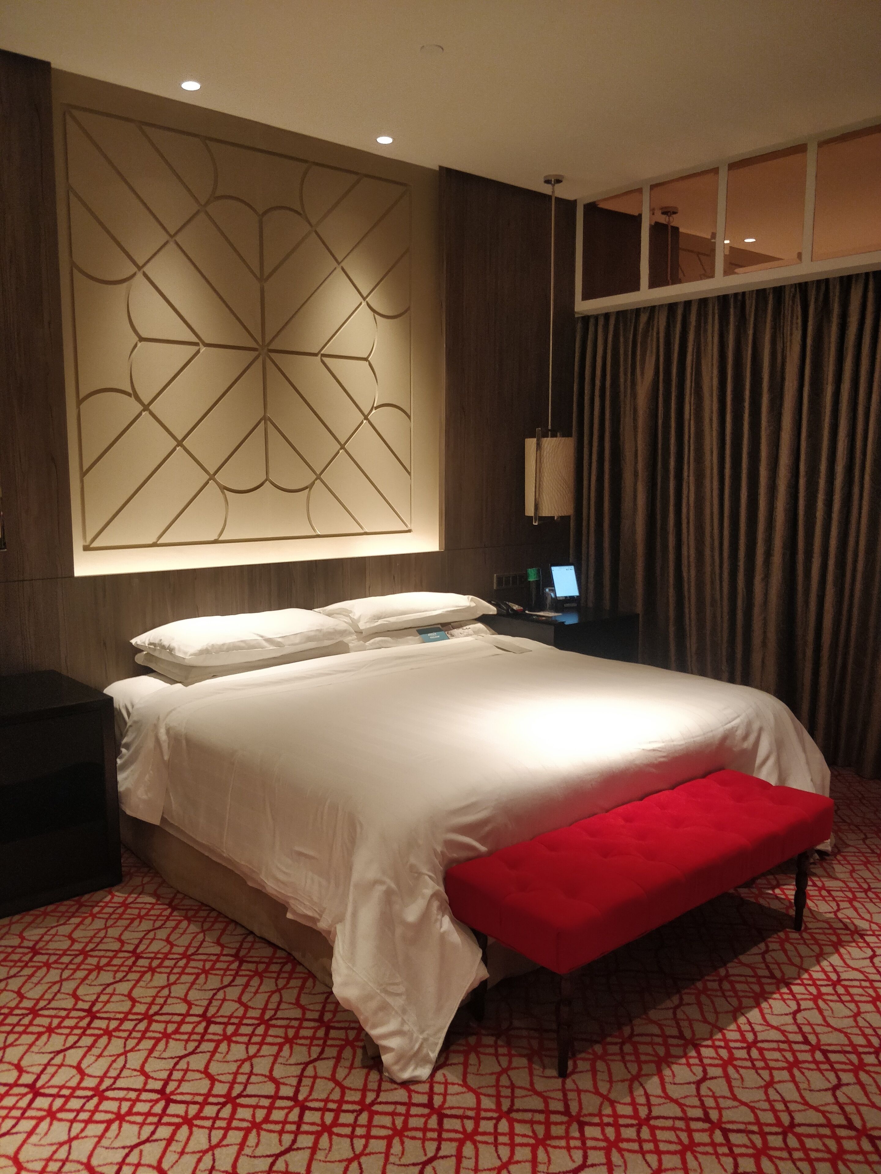 A hotel room with a large bed, a red bench, a wall-mounted light panel, and a window with curtains.