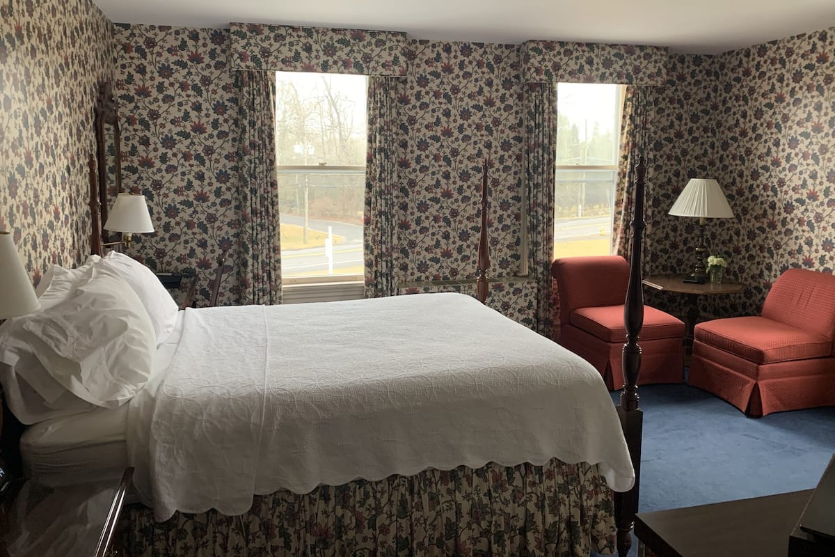 A hotel room with a bed, two chairs, a table, and two windows with curtains.